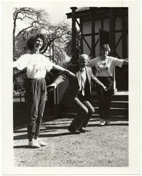 B&W photo of three people conducting in an exercise demonstration on the grounds of a building (which is behind them). In the middle is a man wearing a suit, shirt and tie. On either side of him are two women wearing t-shirts printed with the logo, "Joint Action", and track suit pants.