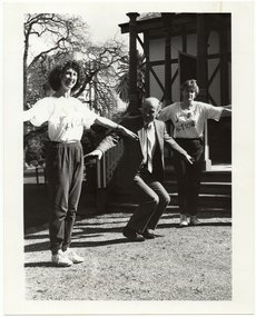 B&W photo of three people conducting in an exercise demonstration on the grounds of a building (which is behind them). In the middle is a man wearing a suit, shirt and tie. On either side of him are two women wearing t-shirts printed with the logo, "Joint Action", and track suit pants.