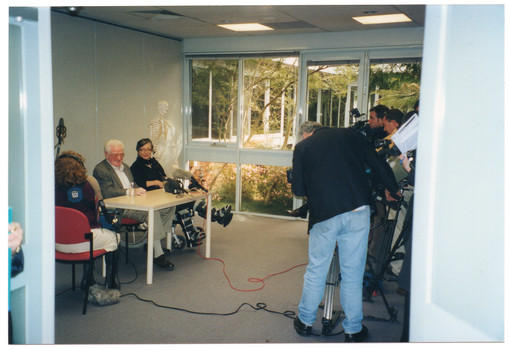 COL photo of a television crew filming a man and a woman who are seated at a table. The woman is a wheelchair user and is wearing leg braces/splints. Behind them, against the wall, is a skeleton on a stand. A crew member is sitting on a chair beside them, holding a boom microphone on a stand. The fluffy boom microphone cover is on the floor, under the chair. Another microphone has been placed on the table. There are two long cables leading from each microphone to the television cameras.