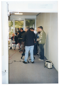 COL photo of a television crew filming. The photo has been taken through an open doorway, with the subjects located in an adjoining room. The crew is standing on the right side of the frame, and their cameras are pointing towards the left of the frame. Beside them is a man sitting on a chair. He is wearing a suit and tie.