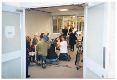 COL photo of a television crew filming a man and a woman who are seated at a table. The crew is kneeling in front of the table to film their subjects at close range. Another crew member is sitting on a chair, recording the audio. She is wearing headphones. Standing beside the table, in front of the windows, are two women, each holding several sheets of paper. Against the wall, there is a skeleton on a stand. In the foreground, there is a tripod camera stand. Although obscured, there are two other people also in the frame.