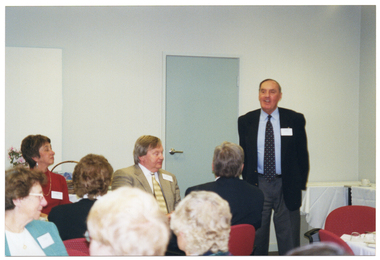 COL photo of a man standing in a room, with people seated at tables in front of him. Behind him, there are two rectangular tables, covered in white tablecloths, pushed against the wall in the corner.