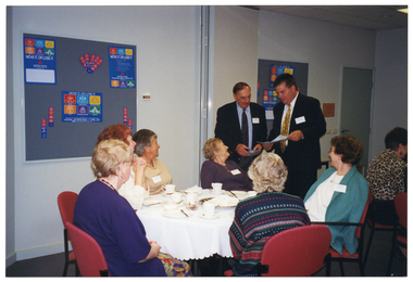 COL photo of two men standing next to a table, at which several women are seated. One man is holding some sheets of paper, while the other man is holding a video cassette. One of the men is speaking, while the other man is looking down. The women are looking at the men. On the table, there are cups and saucers, cake plates and a large platter.