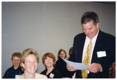COL photo of a man, standing in a room, with several people seated around him. He is looking down at a sheet of paper in his hands. Some of the people seated at the table are looking directly at the camera, smiling.