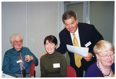 COL photo of a man, standing, as he leans forward towards three people who are seated at a table. He has  one hand placed on the shoulder of the woman in the middle. They are both smiling broadly.