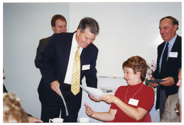 COL photo of a man, standing, as he presents a woman with a certificate. She is seated at a table with a few other people (partially obscured). Two other people can be seen standing beside and behind them.