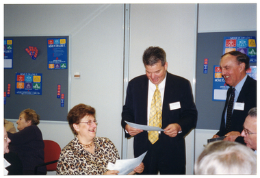 COL photo of two men standing near a woman who is seated at a table with several others. One of the men is looking down at a piece of paper, which he is holding in his hand. The woman also has a piece of paper in her hand. The other man is holding a video cassette in his hand. They are all smiling. In the background, there are some people seated at another table.