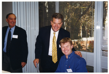 COL photo of a man (standing) as he leans forward to pose for a photo with another man, who is seated at a table. Standing near them is another man. They are all smiling. Behind them are some large windows with thin venetian blinds. Outside the windows is a courtyard with various plants.