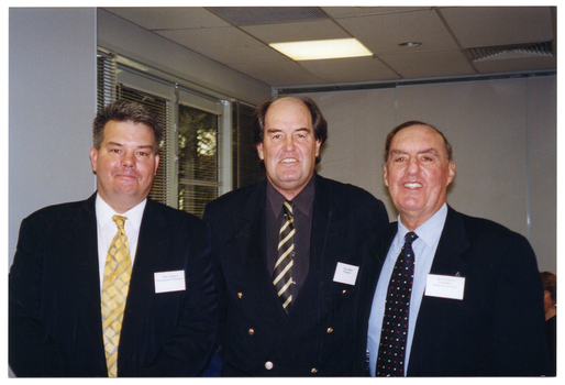 COL photo of three men posing for a photo. They are in a large room. In the background, there is a set of windows with thin venetian blinds to the left. There is a view of some trees through the windows.