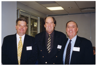 COL photo of three men posing for a photo. They are in a large room. In the background, there is a set of windows with thin venetian blinds to the left. There is a view of some trees through the windows.