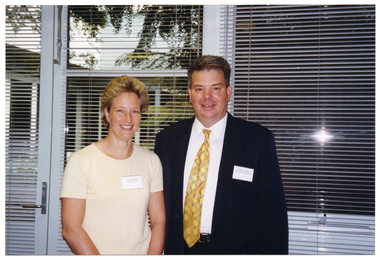 COL photo of two people posing for a photo. They are standing in front of a set of large windows with thin venetian blinds. Through the windows, there is a view of some trees in a courtyard.