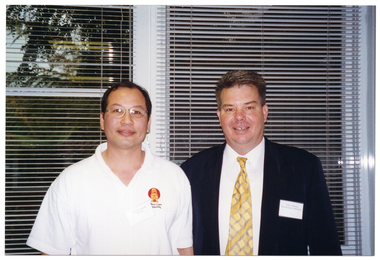 COL photo of two men posing for a photo. They are standing in front of some large windows. The windows have thin venetian blinds. Through the windows, there is a view of some tall trees in a courtyard.