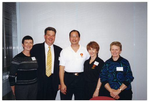 COL photo of a group of five people posing for a photo together. They are standing in a row in front of a wall of closed bi-folding door panels. To the left of the frame, there is a set of windows with thin venetian blinds.