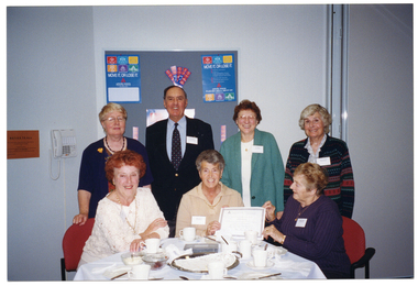 COL photo of seven people, (three seated at a table and four standing behind them), posing for a photo. Two women, seated, hold a certificate up towards the camera. On the table, there are cups and saucers, cake plates, and a large stainless steel platter with a paper doily on top. In the background, there is a large display board with two "Move It or Lose It" posters and various other items pinned to the board.