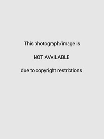 B&W photo of two women posing for a photo. One of the women is passing a trophy to the other woman. She also holding some papers in one hand. Behind them is a cork noticeboard on a wall, to which a graph on a large piece of paper has been pinned. Below the graph is the text, "TEN YEAR"... (partially obscured). There is a dried flower arrangement in a cane basket in front of the wall, beneath the noticeboard.