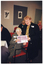 COL photo of two women, (one standing, one seated at a table with others), holding an award certificate together as they pose for a photo. Another person (partially obscured) is sitting next to the woman at the table. In the background, there is another group of people seated at another table. There are some black and white ink paintings on the wall.