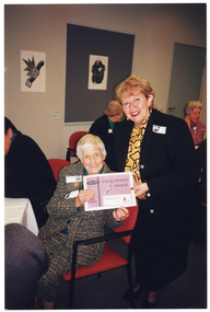 COL photo of two women, (one standing, one seated at a table with others), holding an award certificate together as they pose for a photo. Another person (partially obscured) is sitting next to the woman at the table. In the background, there is another group of people seated at another table. There are some black and white ink paintings on the wall.