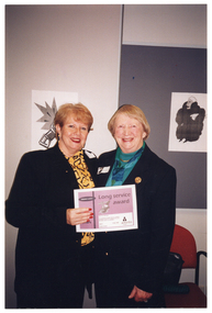 COL photo of two women, both standing, posing for a photo. One of the women is holding up a certificate. On the certificate is the text, "Long Service Award". Behind them are two unframed black and white pictures, one on the wall and one on a display board.