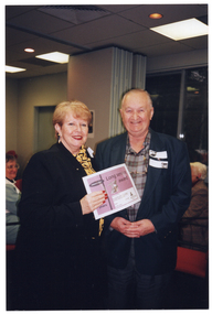 COL photo of two people, both standing, posing for a photo together. The woman is holding up a certificate. On the certificate is the text, "Long Service Award". In the background, groups of people are seated at various tables.