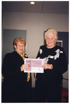 COL photo of two women, both standing, posing for a photo. Together, they are holding up a certificate. On the certificate is the text, "Long Service Award". In the background, there is a closed door and an unframed black and white picture on a display board.