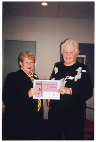 COL photo of two women, both standing, posing for a photo. Together, they are holding up a certificate. On the certificate is the text, "Long Service Award". In the background, there is a closed door and an unframed black and white picture on a display board.