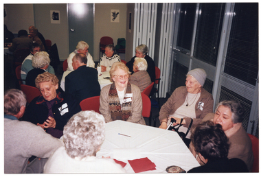 COL photo of several small groups of people seated at octagonal tables positioned around a large room. They are chatting and smiling. On one side of the room, there is a large set of windows with thin venetian blinds. On the same side, there is a set of bi-fold doors which have been opened and folded back to join two rooms together. There are some black and white ink paintings on the wall.