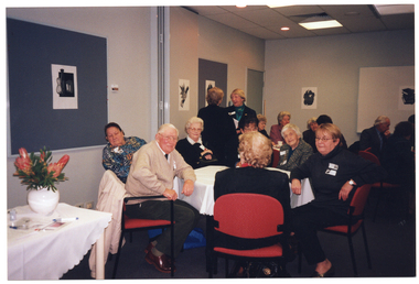COL photo of several groups of people seated at octagonal tables in a large room. They are chatting and smiling. Two people are standing, facing one another. There is a bouquet of proteas in a white vase on a rectangular table against the wall. There are several unframed black and white ink paintings adorning the walls.