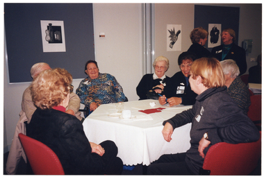 COL photo of a group of people seated at an octagonal table in a large room. They are chatting and smiling. In the background, two other people are standing, facing one another. There are some black and white ink paintings on the walls.