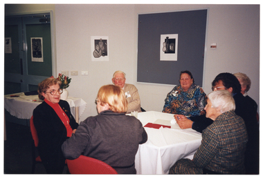 COL photo of a group of people seated at an octagonal table in a large room. A few people are facing the camera, while the others have been Photographed from behind. There are some black and white ink paintings on the doors and walls.