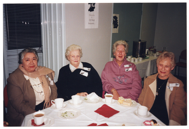 COL photo of four women seated at an octagonal table. There are cups of tea or coffee and small plates of cake on the table. In the background, there is a long rectangular table against the wall. On the table is a large stainless steel urn, several milk jugs, glasses and glass carafes of water, and other miscellaneous items. On the wall, there is an unframed black and white ink painting, and another on a bi-fold door (which has been folded back). Below the picture, there is the text, "Welcome to all Op Shop treasure...".