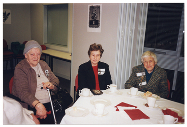 COL photo of three women seated at an octagonal table. One woman is holding a walking stick. There are cups of tea or coffee and plates of cake on the table. Behind them is a large set of windows with thin venetian blinds and a set of bi-fold doors, which have been folded back. On the outermost bi-fold door panel, there is an unframed black and white ink painting, and another on an adjoinging wall. In the background, there are two closed servery doors set into the wall. There is a narrow rectangular table below the servery.