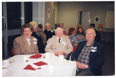 COL photo of three people seated at an octagonal table, smiling. There are cups of tea or coffee and small plates on the table. Behind them is a large set of windows with thin venetian blinds and a set of bi-fold doors, which have been folded back. There is an unframed black and white picture on the outermost bi-fold door panel. In the background, there is a large stainless steel urn on a table. There is another group of people seated at another table behind them. There are two unframed black and white pictures on the walls. There is a large whiteboard which has been affixed to one of the walls and, below it, a vase of flowers on a narrow rectangular table.