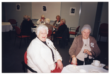 COL photo of two women, seated at a table, posing for a photo. One of the women is holding a walking stick. There are cups of tea or coffee and small plates with cake on the table. In the background, another group of women are seated at another table.