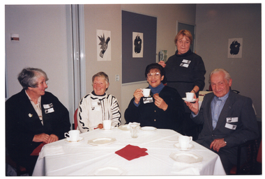 COL photo of five people gathered around an octagonal table: four of them are seated, while a fifth person is standing behind one of them. There are cups and small plates on the table. Two people are holding their cups and saucers up. There are three black and white ink paintings on the walls, one of which is on a display board. One of the paintings is on the outermost panel of a set of bi-fold doors, which have been folded back.