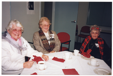 COL photo of three women seated at a table. They are smiling. On the table, there are cups of tea or coffee and small plates. In the background, there is a black and white ink painting on the wall.