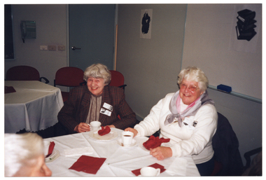 COL photo of two women seated at a table. They are facing the camera and smiling. Facing them is another person, also seated at the table, but only partially visible in the frame. On the table, there are cups of tea or coffee and small plates. In the background, there is a telephone affixed to the wall, and two black and white ink paintings on the wall.