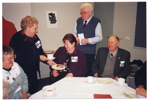 COL photo of several people around a table: three are seated and two are standing. One woman, standing, is passing a cup and saucer and a small plate of cakes to another woman, who is seated. There are several other cups and saucers, and small plates on the table. In the background, one person (standing) is facing the wall and has been from behind. There is a black and white picture on the wall. Two other people, who are also seated at the table, are partially obscured and mostly out of frame.