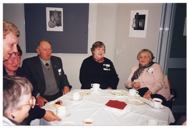 COL photo of several people seated at an octagonal table. Three people are on the edge of the frame, partically obscured. One of them is a woman who is standing and leaning forward. She is holding a cup and saucer above the table. On the table, there are several cups and saucers, and small plates.