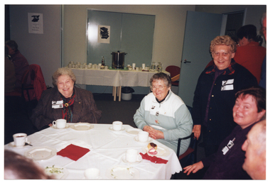 COL photo of several people gathered around a table. One person is standing, while the others (two of whom are partically obscured) are seated at the table. In the foreground, to the right, there are a few people walking behind them, away from the camera. On the table, there are several cups and saucers, and small plates. In the background, there are a few people are seated at a table to the left while, in the centre, there is a long table with cups and saucers, an urn, and carafes of water.