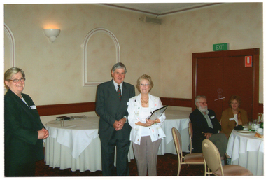 COL photo of a man and a woman posing for a photo. The woman is holding a framed award. They are standing in front of some round tables at a reception centre. To their right is another woman, standing. Her face is turned towards the camera and she is smiling. To their left are two people seated at a round table.