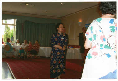 COL photo of a woman approaching a few people standing to the right of the frame, in the foreground. She is holding a piece of paper in her hand. Another woman is standing on a low dais, covered in red fabric. In the background, there are several people seated at a large round table.