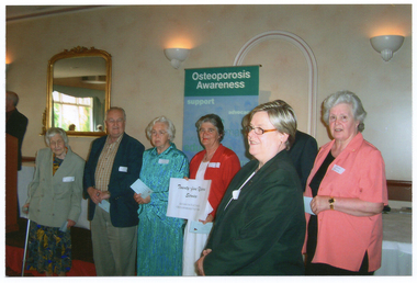 COL photo of a small group of people standing in a row, each holding a small piece of paper. One woman has a walking stick, while another woman is holding up a sign that reads "Twenty-Five Years Service", "Arthritis Victoria", "Osteoporosis Victoria". Standing in front of them is another woman. Behind them is a banner with the heading "Osteoporosis Awareness", with various text below it, including 'support' and 'advocacy'.