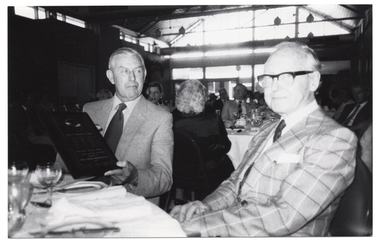 B&W photo of two men sitting at a large round table. On the table, there are several wine glasses, and plates with cutlery. One of the men is holding a framed award. In the background, groups of people are seated at several other tables.