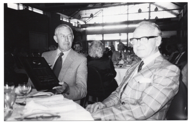 B&W photo of two men sitting at a large round table. On the table, there are several wine glasses, and plates with cutlery. One of the men is holding a framed award. In the background, groups of people are seated at several other tables.