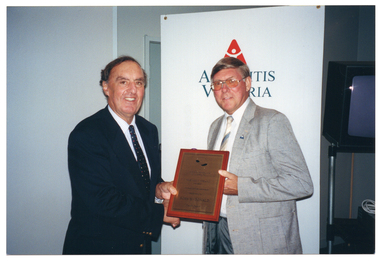 COL photo of two men standing side by side, holding a framed award between them. Behind them is a tall vertical freestanding banner containing the text and logo for Arthritis Victoria (partially obscured). To one side the banner, there is a television set on a stand.