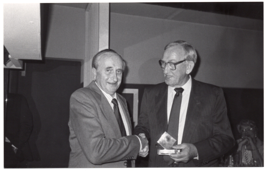 B&W photo of two men shaking hands. One of the men is holding a small award trophy in his other hand. In the background, there is a woman who is seated.