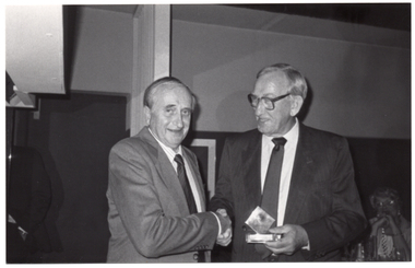 B&W photo of two men shaking hands. One of the men is holding a small award trophy in his other hand. In the background, there is a woman who is seated.