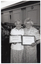 B&W photo of two women, standing close together, each holding a framed award certificate. They are in a large hall. There are several other people standing in the background.