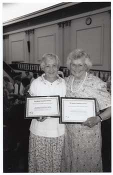 B&W photo of two women, standing close together, each holding a framed award certificate. They are in a large hall. There are several other people standing in the background.