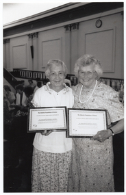 B&W photo of two women, standing close together, each holding a framed award certificate. They are in a large hall. There are several other people standing in the background.